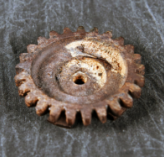 A small, corroded, gear-shaped metal object, likely a mechanical part recovered from an archaeological or forensic site.