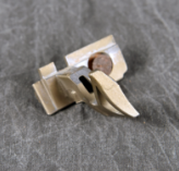 A small, complex, tan-colored plastic or metal part, possibly a latch or clip mechanism, with a metal pin inserted through it, displayed on a dark fabric background.