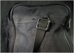 A close-up view of a black bag, showing the attachment point of a black woven shoulder strap to a plastic buckle.