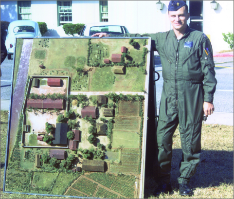 A man in a flight suit stands proudly next to a large, detailed topographical model of the Son Tay POW camp, used for planning the rescue mission.