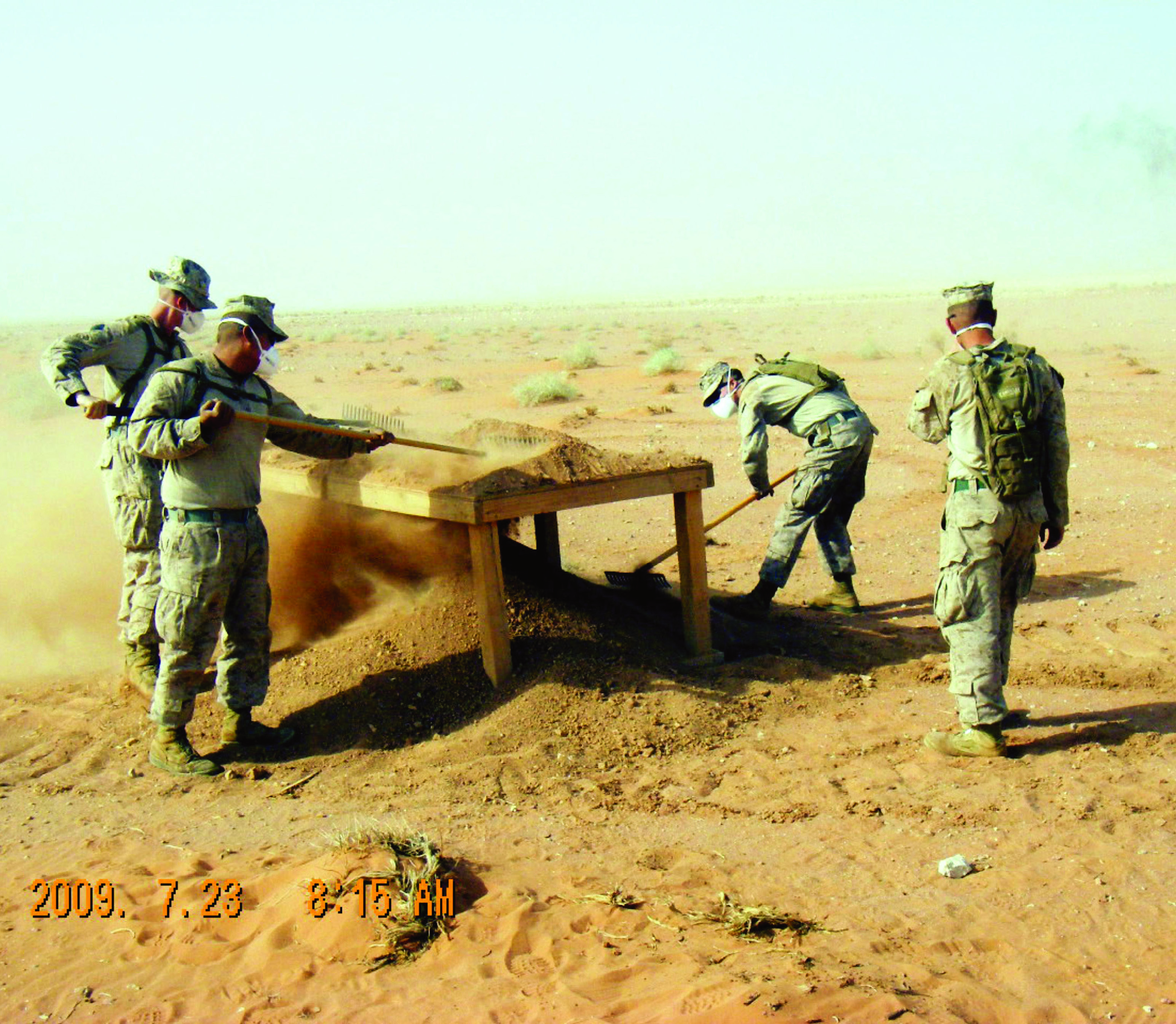 Marines in full combat gear and dust masks sift sand through a large screen at a recovery site in the desert.
