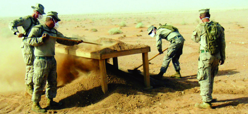 Marines in full combat gear and dust masks sift sand through a large screen at a recovery site in the desert.