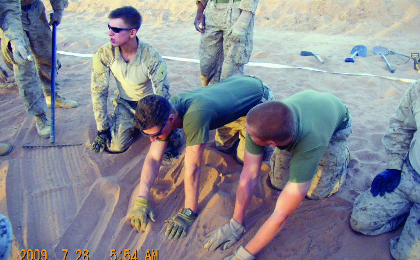 Marines conduct a meticulous search by hand, crawling and smoothing the sand in a designated lane at a recovery site, dated 28 July 2009.