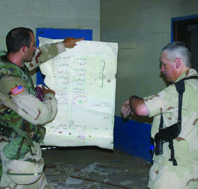 Two U.S. soldiers in a damaged room, one pointing to details on a large, captured Arabic map or document.