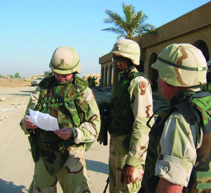 Three soldiers in desert combat gear stand in a street in a Middle Eastern village, reviewing a document.