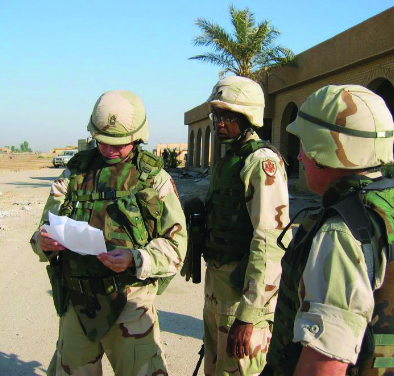 Three soldiers in desert combat gear stand in a street in a Middle Eastern village, reviewing a document.