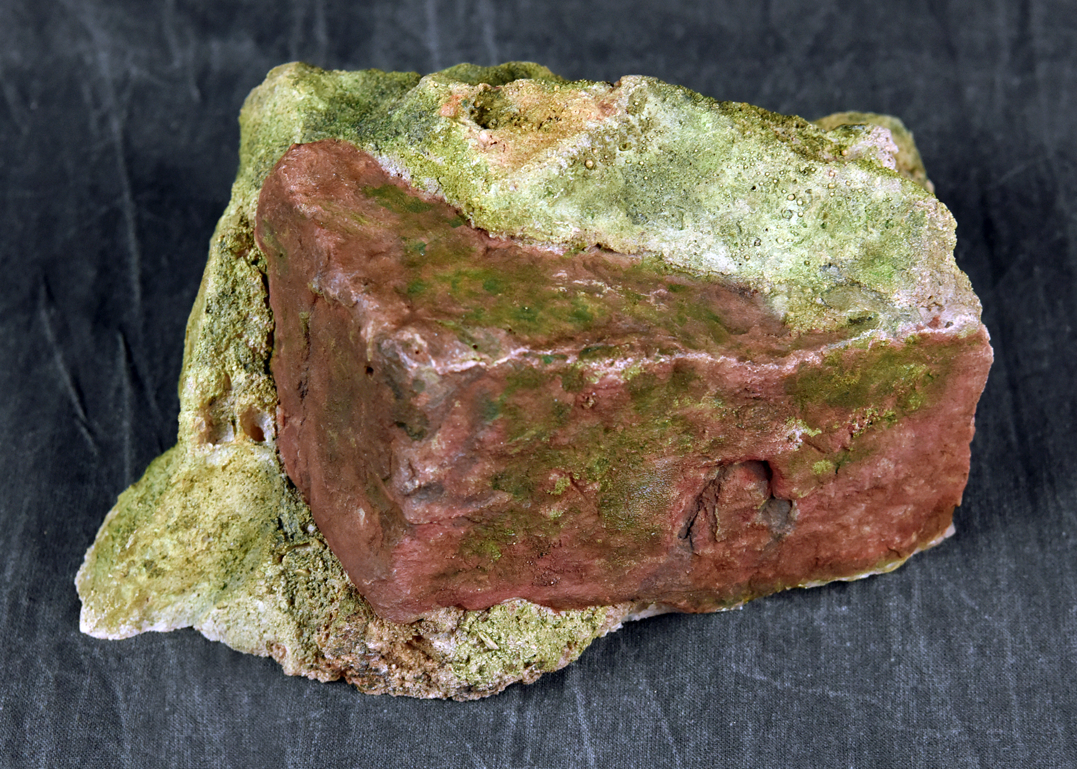 A single piece of composite rock against a black background, featuring a reddish, block-like section embedded in a porous, greenish-yellow mineral formation.
