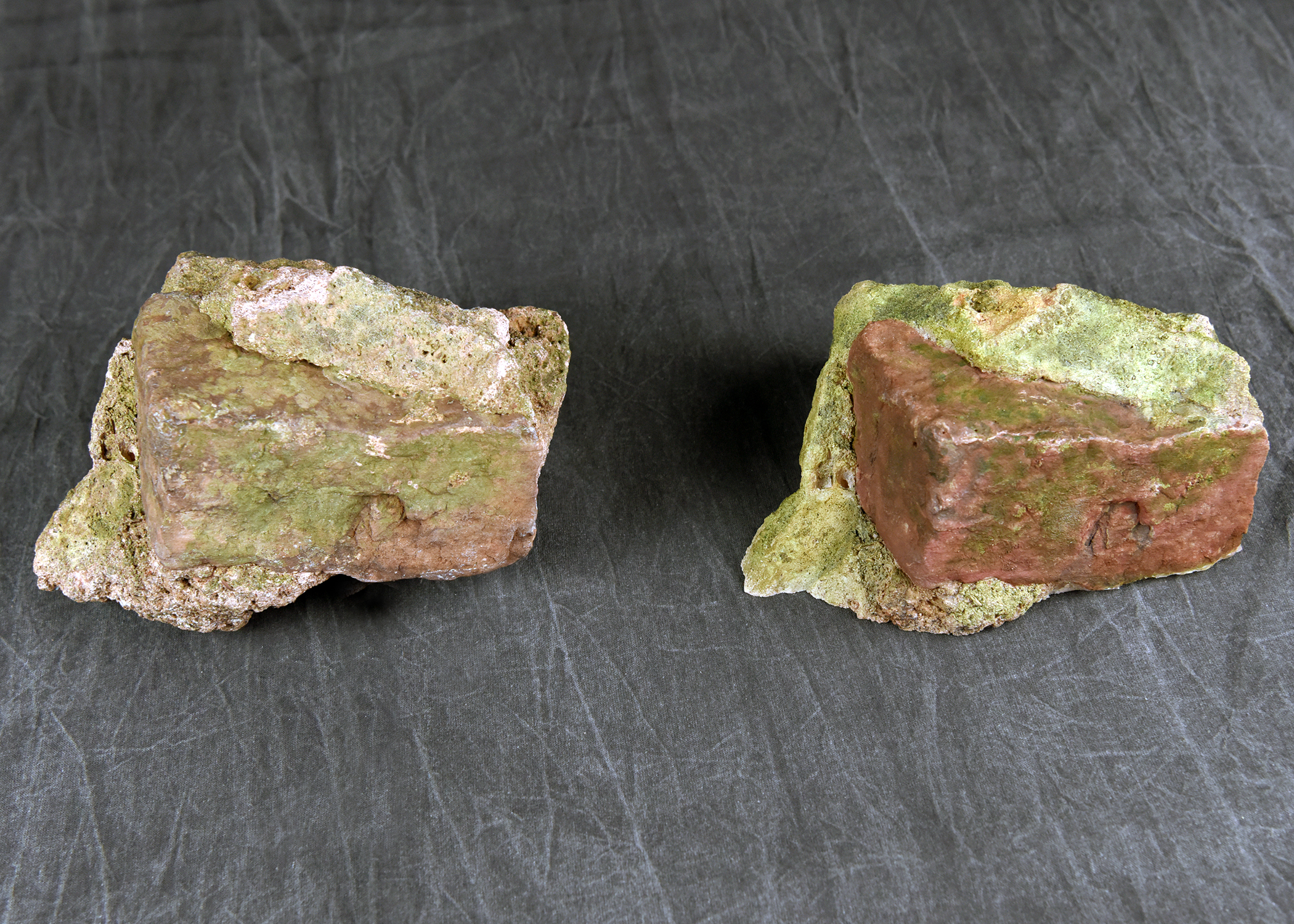 Two pieces of irregularly shaped, rock-like material with reddish-brown and greenish-yellow coloration, resting on a dark surface.