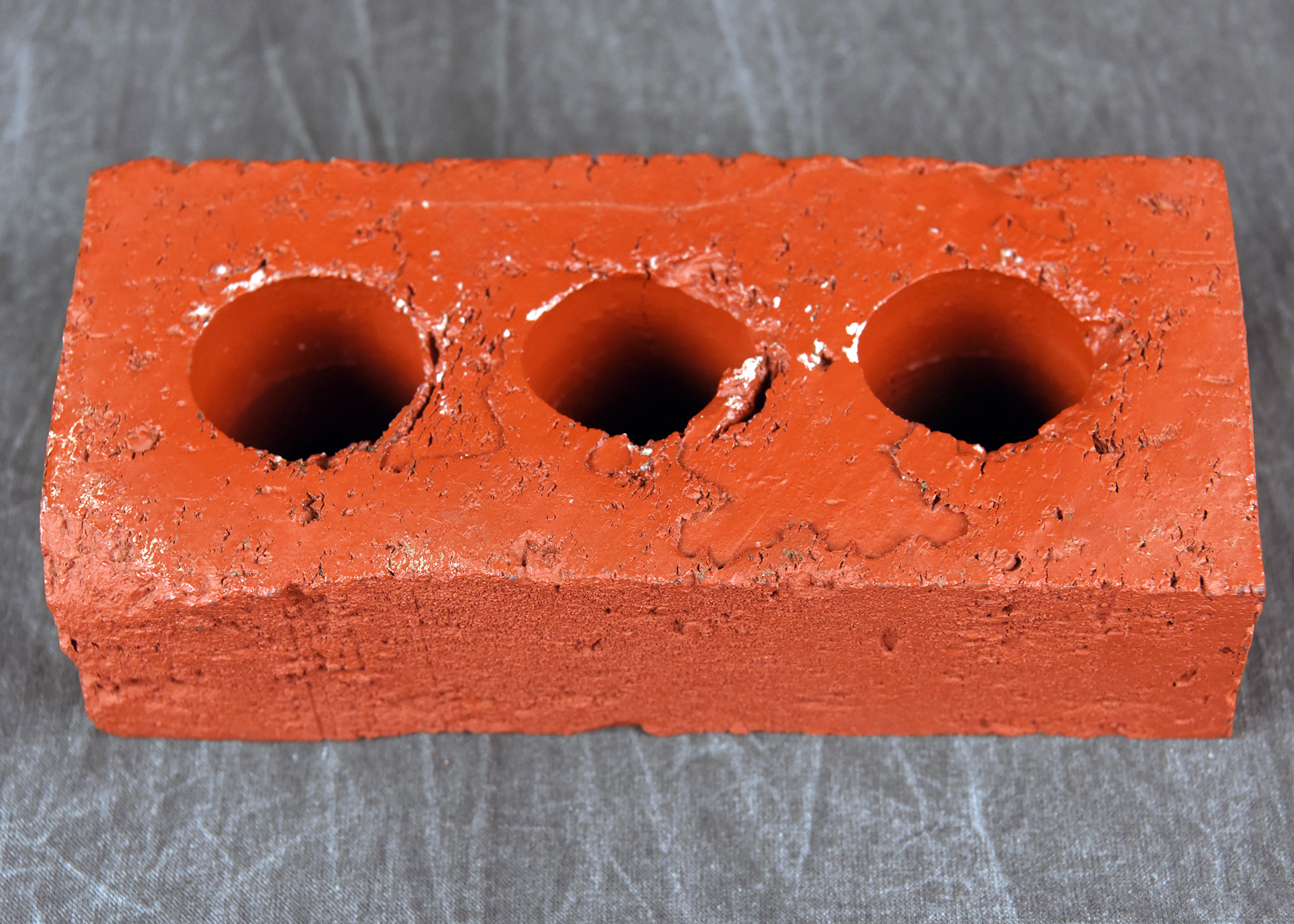 A close-up, high-resolution photograph of a single red brick with a rough texture. Three circular holes run vertically through its center. The brick is resting on a gray, woven fabric background.