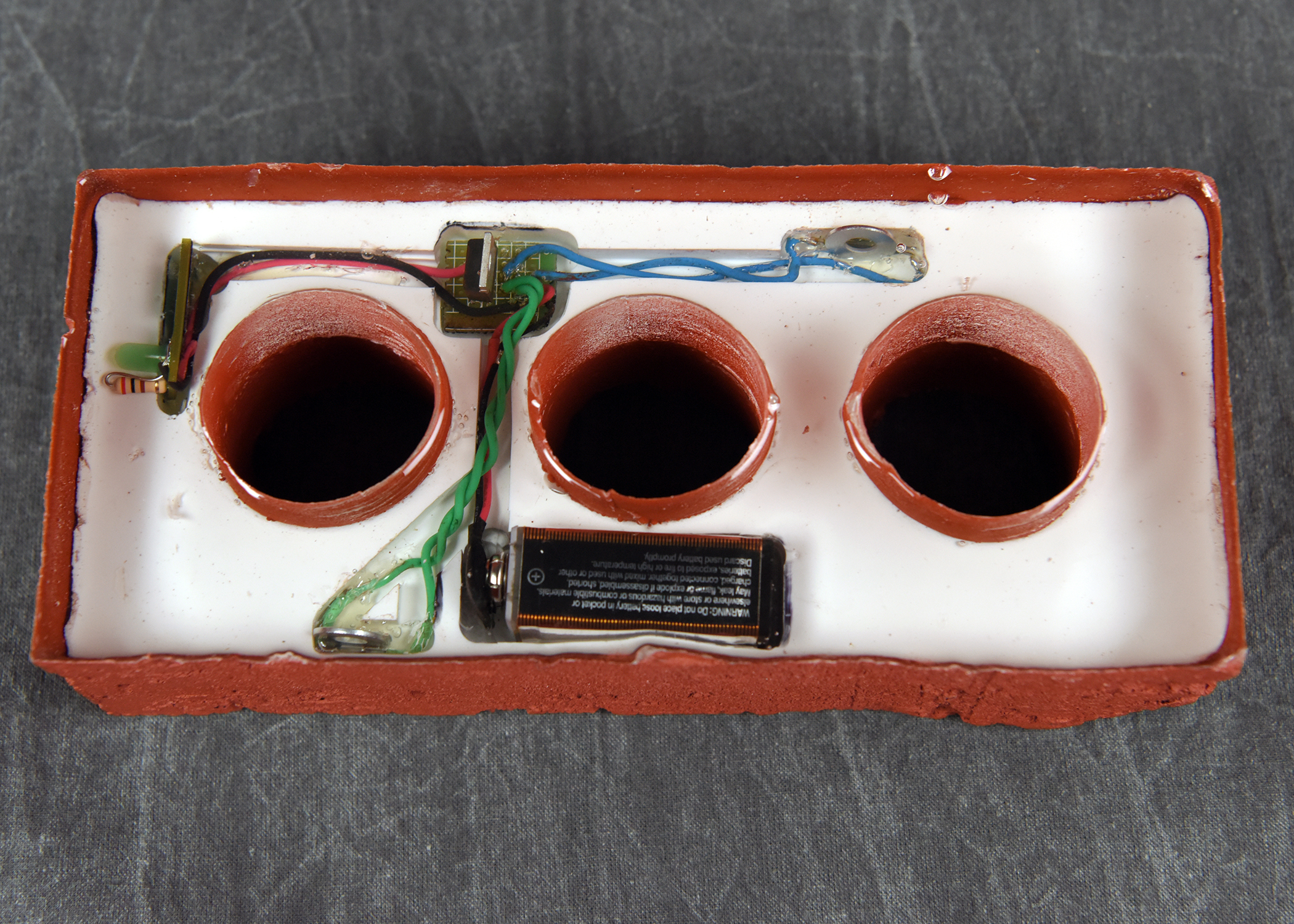 A white rectangular tray with a reddish-brown border, containing three round, dark-colored wells and various electronic components with wiring.