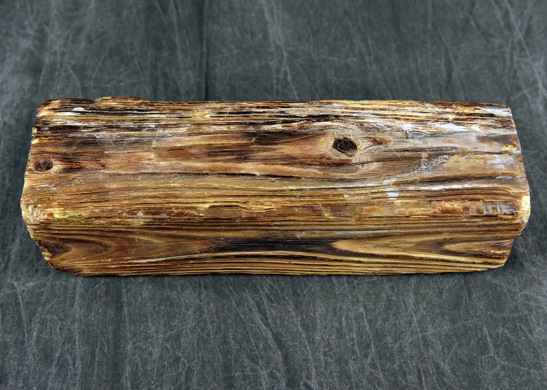 A detailed, high-resolution photograph of a rectangular block of petrified wood. The surface displays a prominent, weathered grain pattern with horizontal streaks of brown, black, and off-white. The block is resting on a gray, woven fabric background.