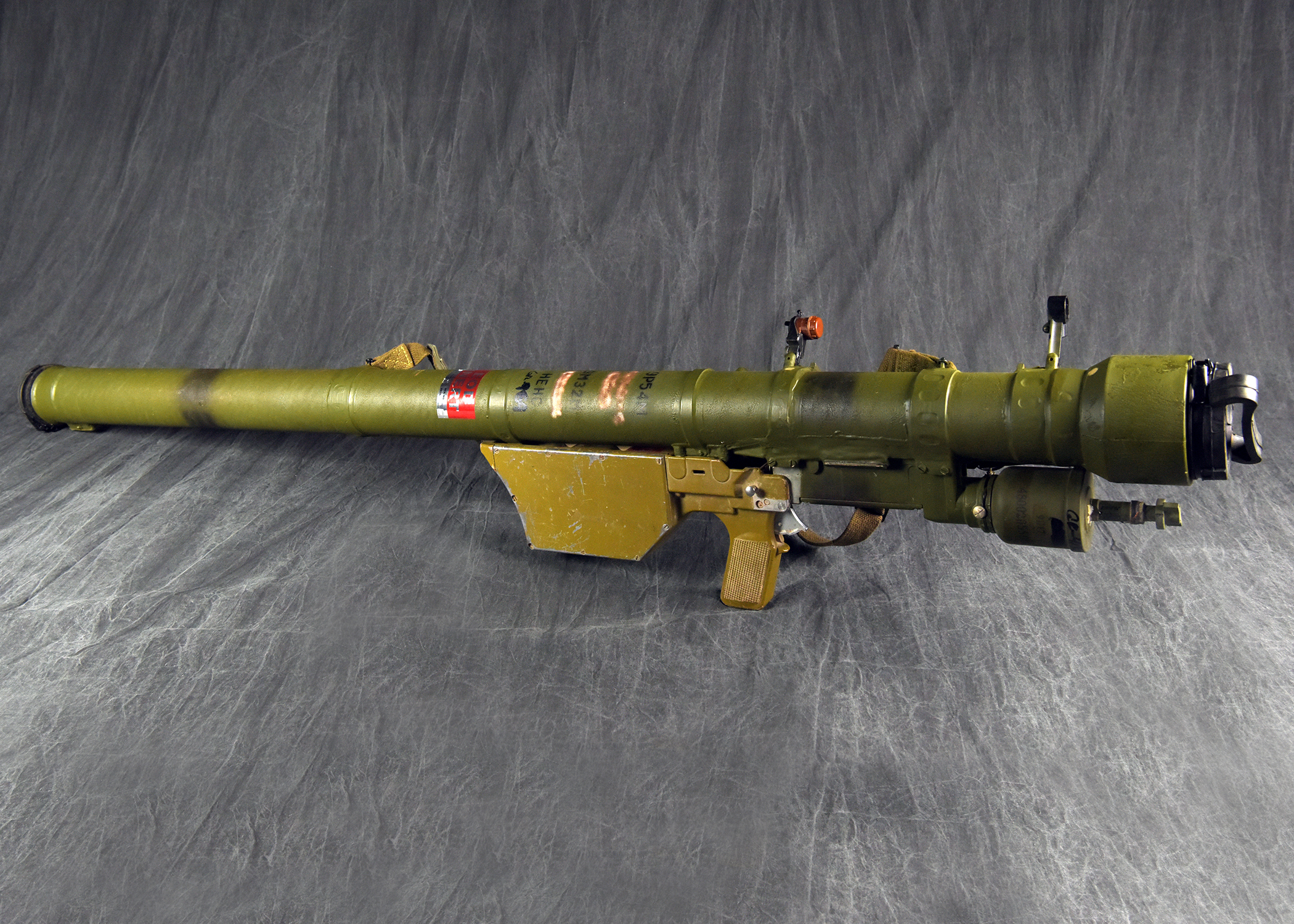 A green, shoulder-fired surface-to-air missile launcher with its gripstock and trigger mechanism is displayed against a dark fabric background.
