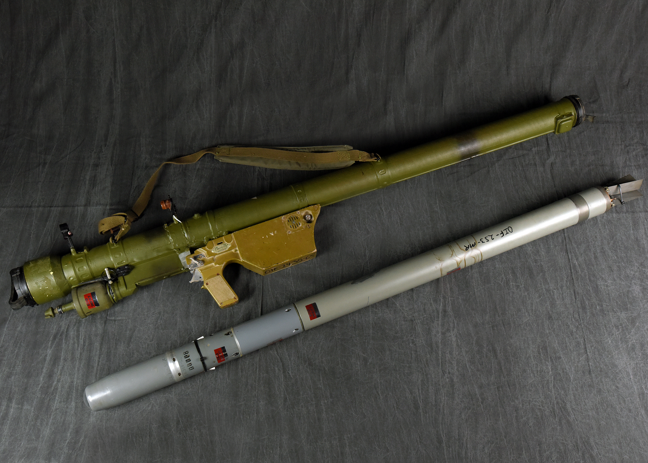A green shoulder-fired missile launcher and a separate gray missile are laid out parallel to each other on a dark fabric background.