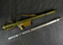 A green shoulder-fired missile launcher and a separate gray missile are laid out parallel to each other on a dark fabric background.