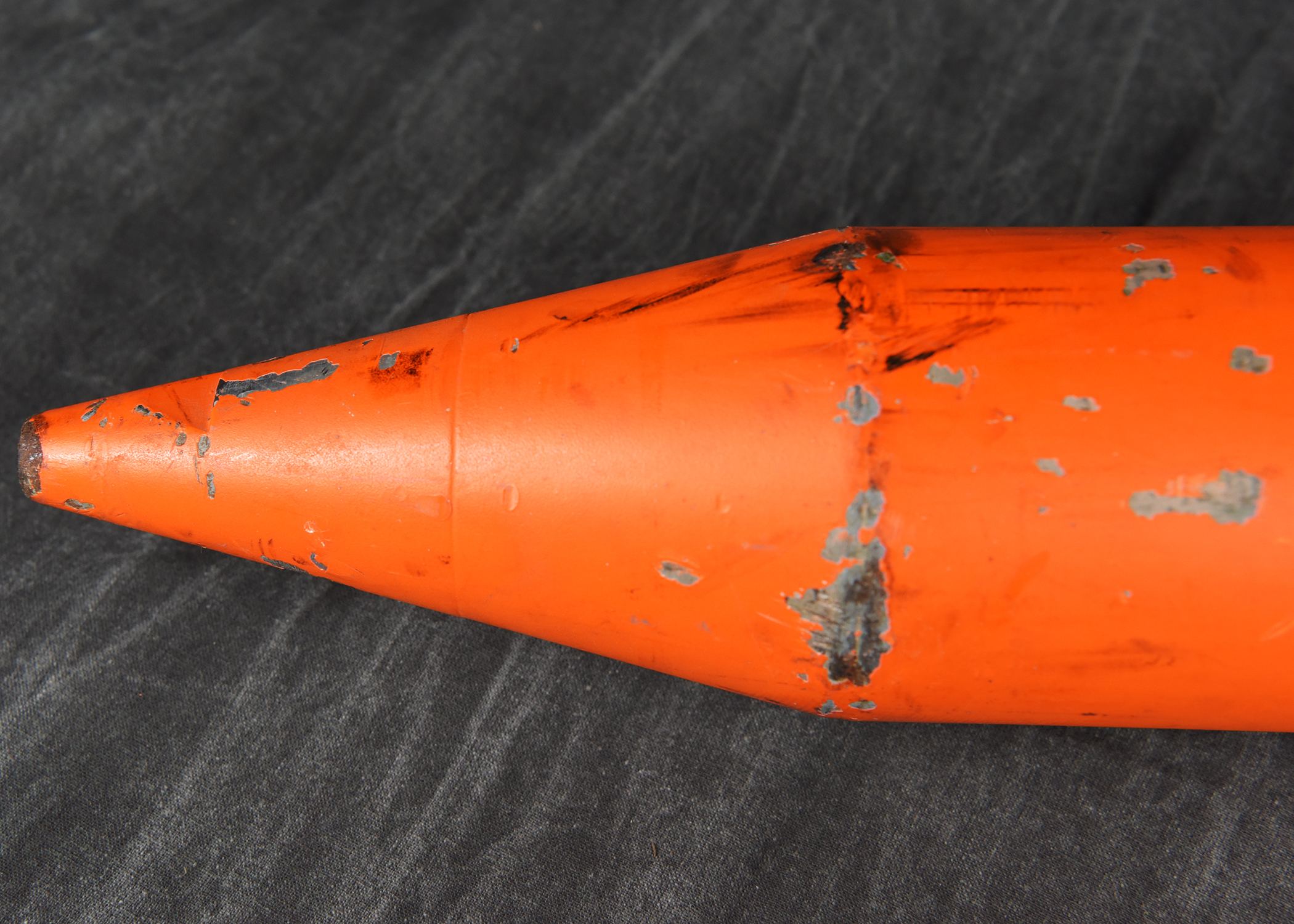 A close-up photograph of the pointed, orange nose cone of a cylindrical instrument. The surface shows some scuff marks and wear. The background is a dark, wrinkled fabric.