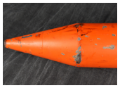 A close-up of the nose cone of an orange munition, showing scuff marks and wear. The device is resting on a dark fabric surface.