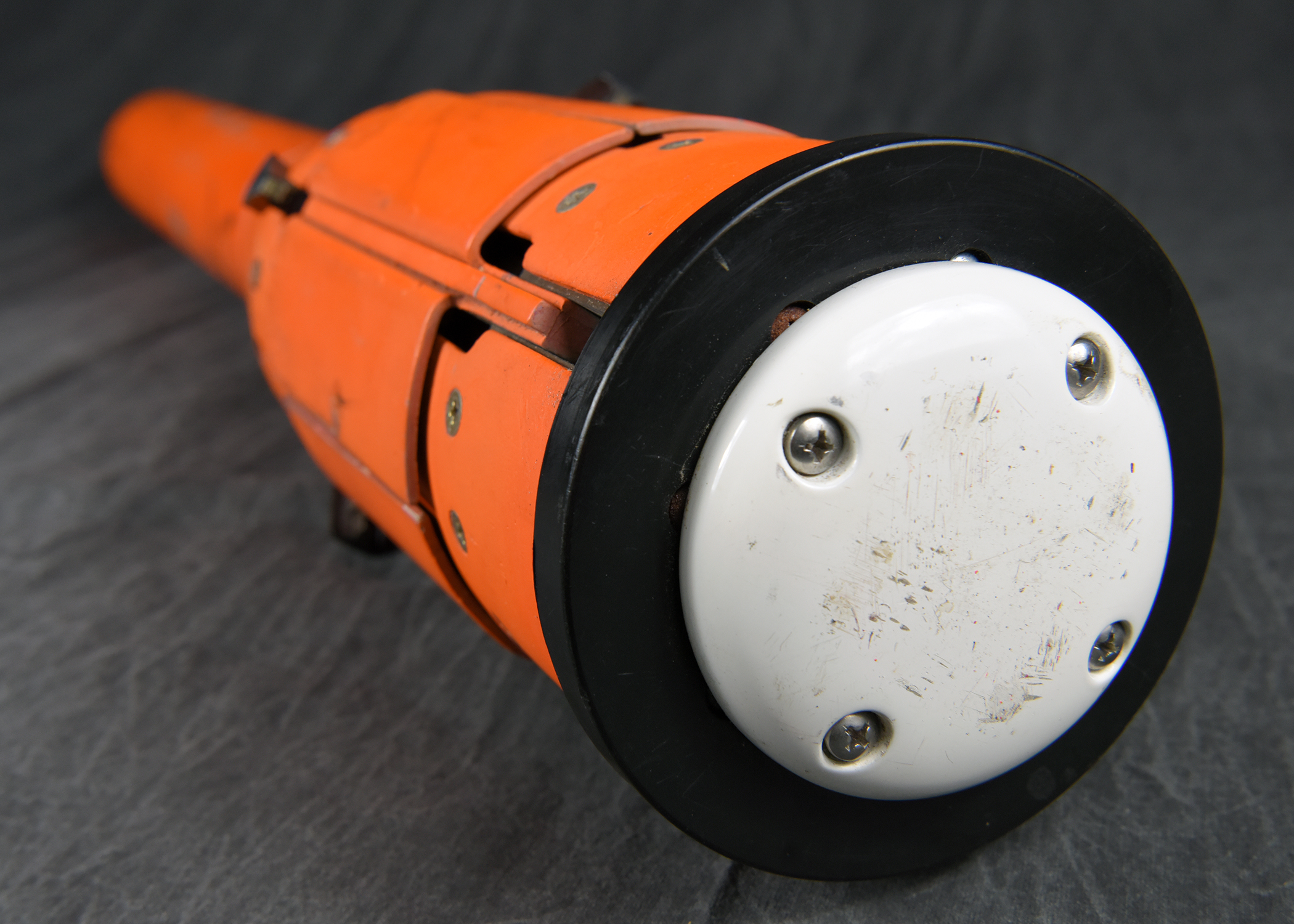The base of an orange, cylindrical instrument. A flat, white end cap is secured with four screws and is surrounded by a black circular bumper. The background is a dark, wrinkled fabric.