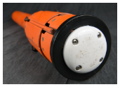 A close-up of the tail end of an orange munition shows a white circular base plate secured with four screws and encircled by a black ring. The device is resting on a dark fabric surface.