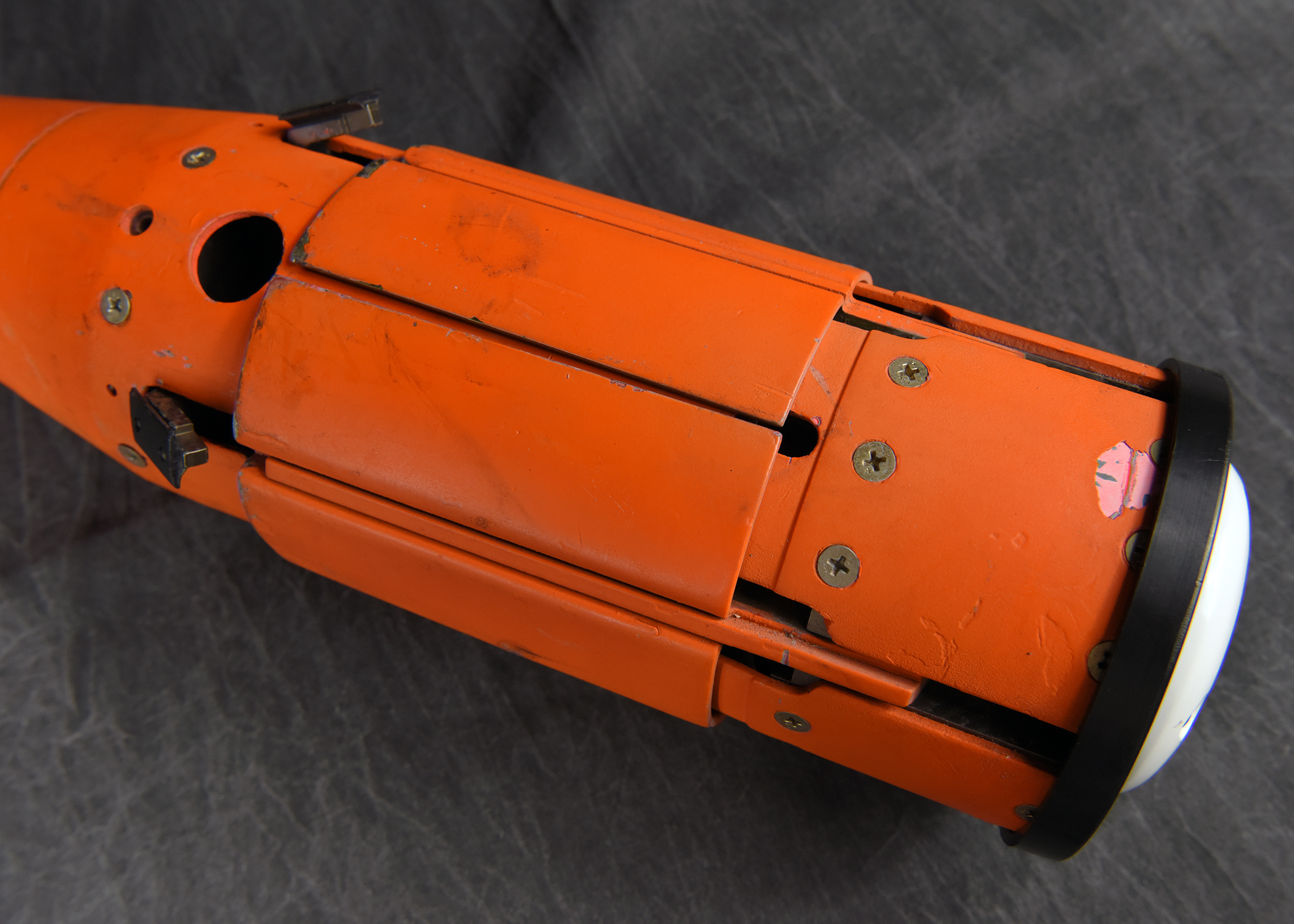 The mid-section of an orange, cylindrical instrument, showing mechanical details such as hinged panels, seams, and a circular port on its side. The background is a dark, wrinkled fabric.