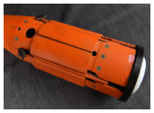 A close-up view of the tail assembly of an orange munition. The fins are shown retracted, folded into slots in the device's body. The munition is resting on a dark, rumpled fabric.