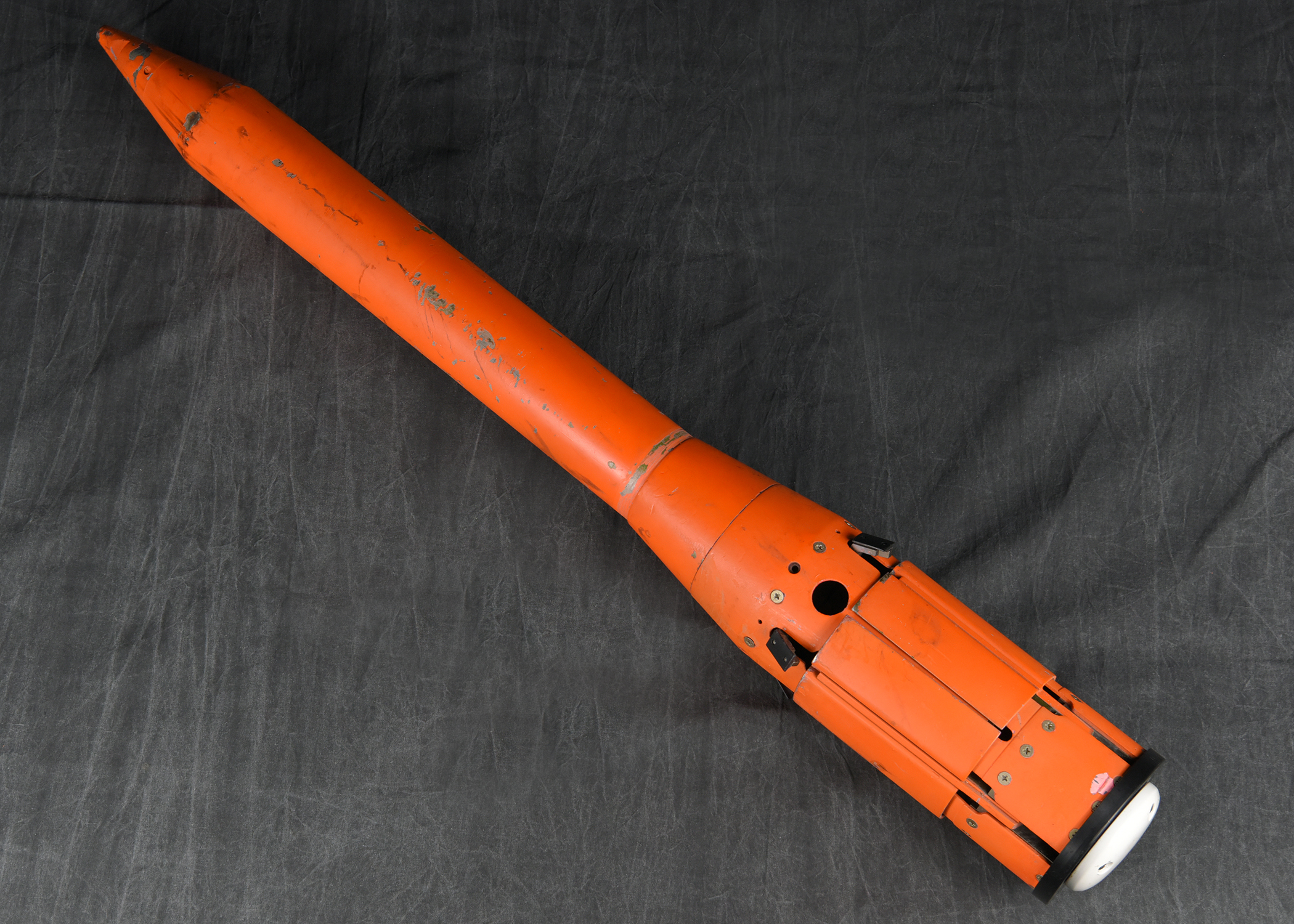 A full-length photograph of a long, orange, probe-like instrument lying on a dark, wrinkled surface. The device has a pointed nose cone at one end and a wider, mechanical base at the other.