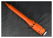 A bright orange, tube-shaped munition rests on a dark, rumpled fabric. The device has a sharply pointed nose and a wider tail assembly, shown with its fins retracted.