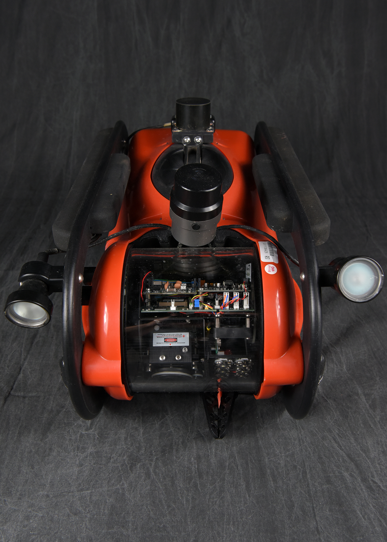 A top-down view looking into the open chassis of an orange submersible device, revealing its internal electronics, including a circuit board and wiring. Two forward-facing lights are visible on either side.A top-down view looking into the open chassis of an orange submersible device, revealing its internal electronics, including a circuit board and wiring. Two forward-facing lights are visible on either side.