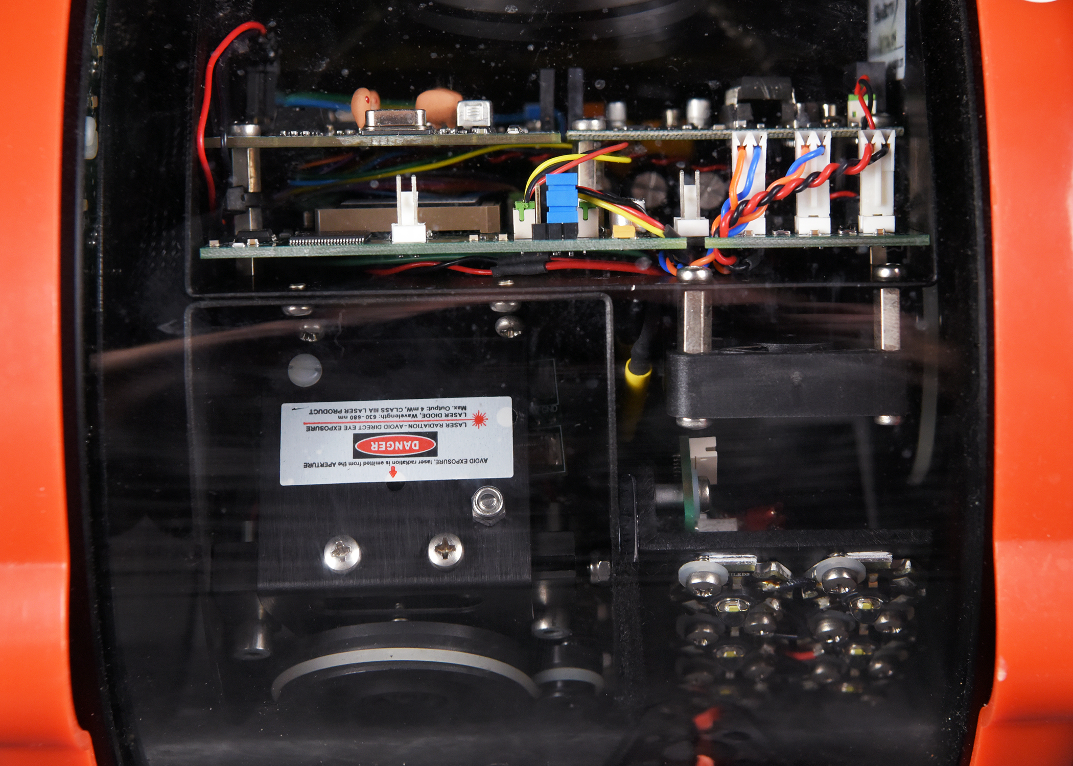 A close-up photograph of the internal electronics of a device. The image shows a complex assembly of circuit boards, multi-colored wires, and a silver warning label indicating a laser danger.