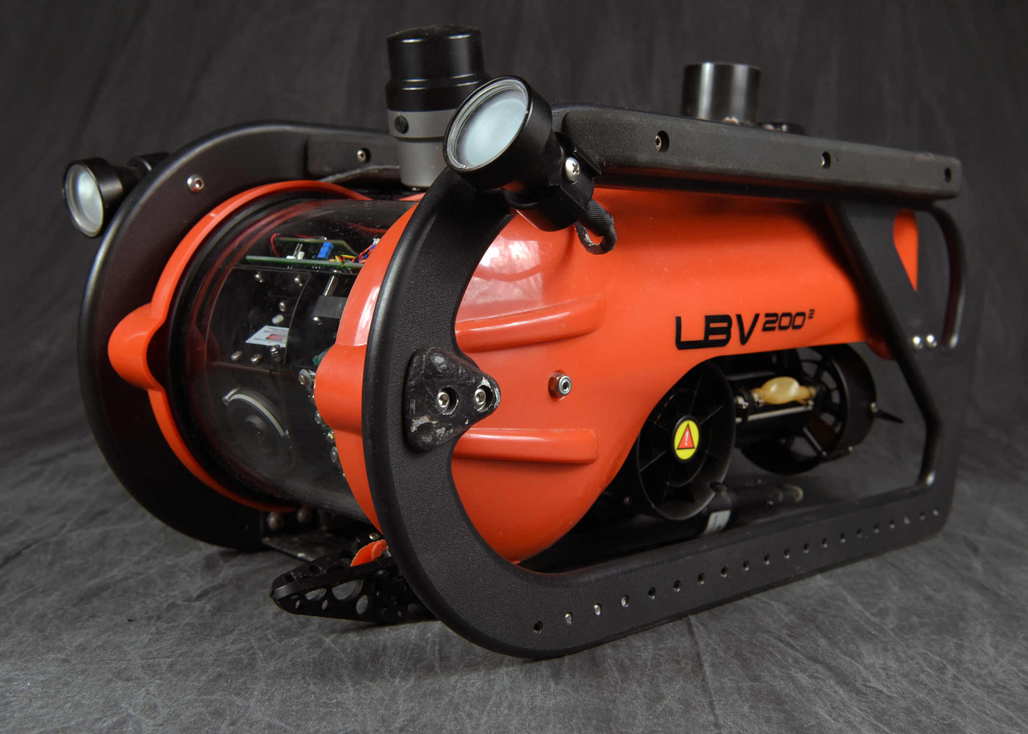A side profile of an orange and black remotely operated vehicle (ROV) on a dark surface. The device has a black protective frame, two forward-facing lights, and the text 'LBV200' printed on its side. A clear dome at the front reveals some of its internal components.