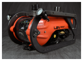 An orange and black LBV200 remotely operated vehicle (ROV) is shown resting on a dark fabric background. It features a protective black frame, external lights, and a clear rear dome that reveals its internal electronics.