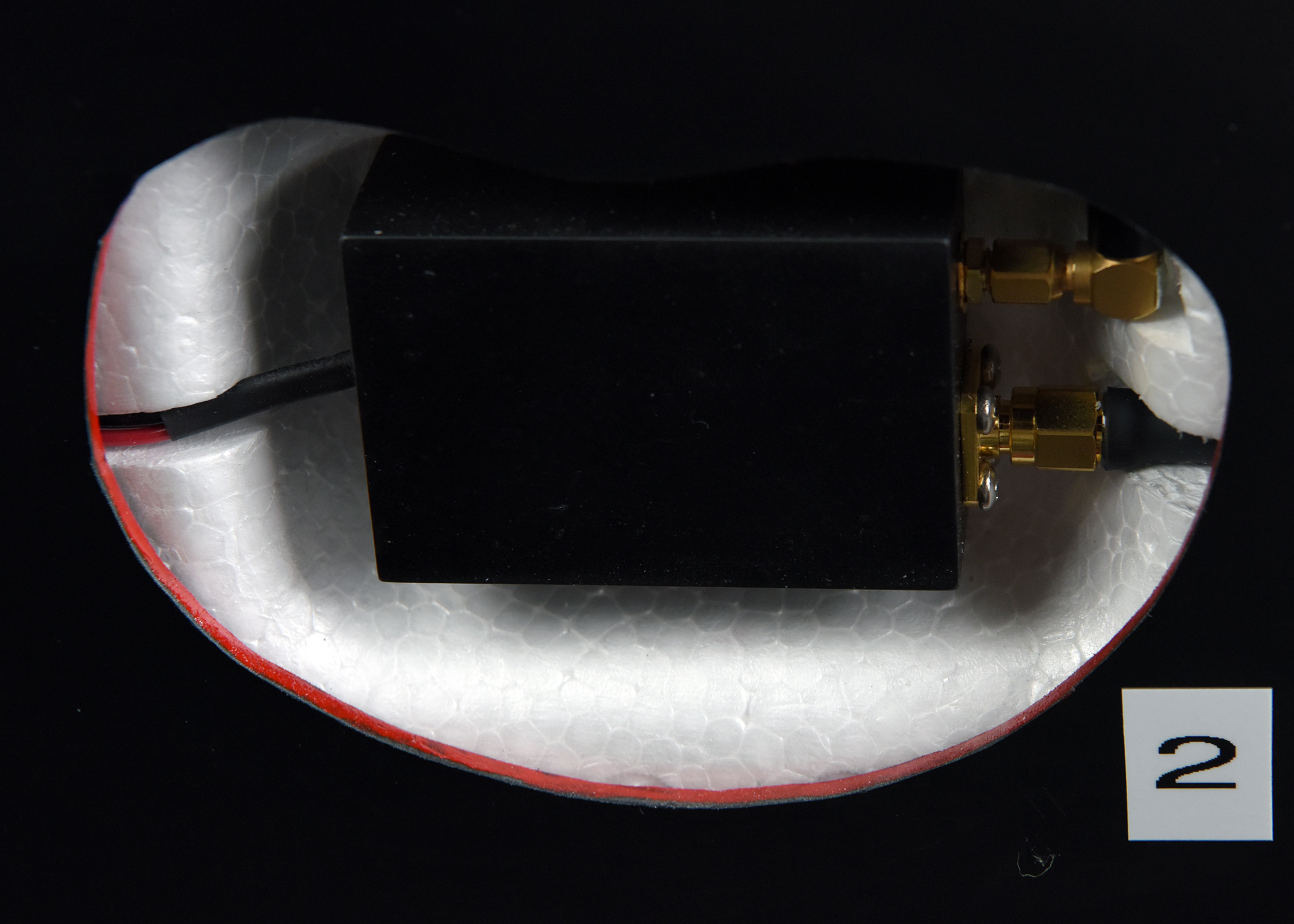 A small, black rectangular electronic component with two gold-colored connectors, viewed through an oval cutout in a white foam-like material. A small label with the number 2 is visible in the corner.