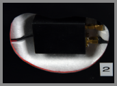 A close-up of a black electronic device with gold-colored connectors, marked as 2, recessed into a hidden compartment within a car bumper.