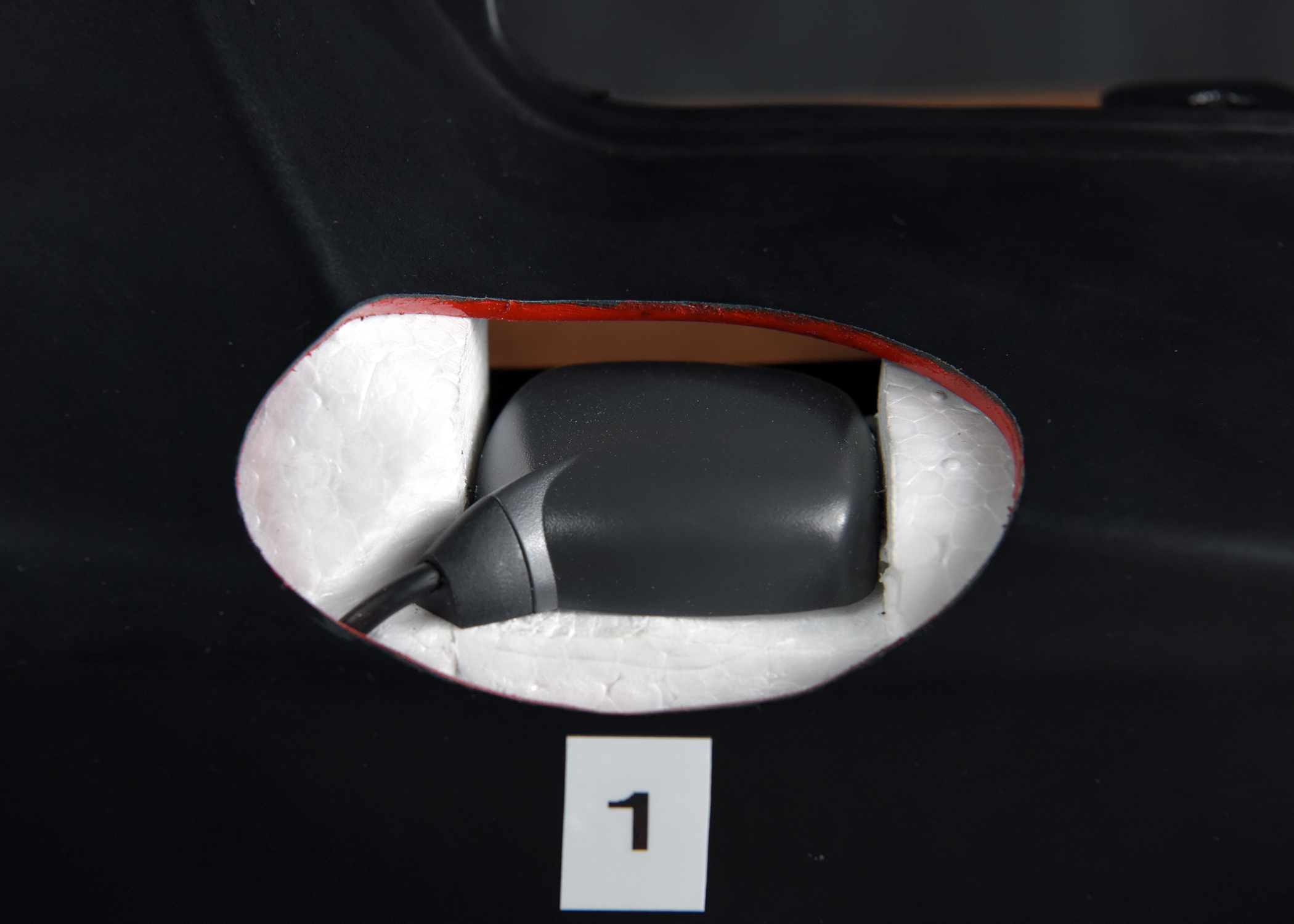 A dark gray, boxy component is seen through an oval opening in a black surface. The opening is lined with white padding, and a small white label with the number 1 is placed below it.
