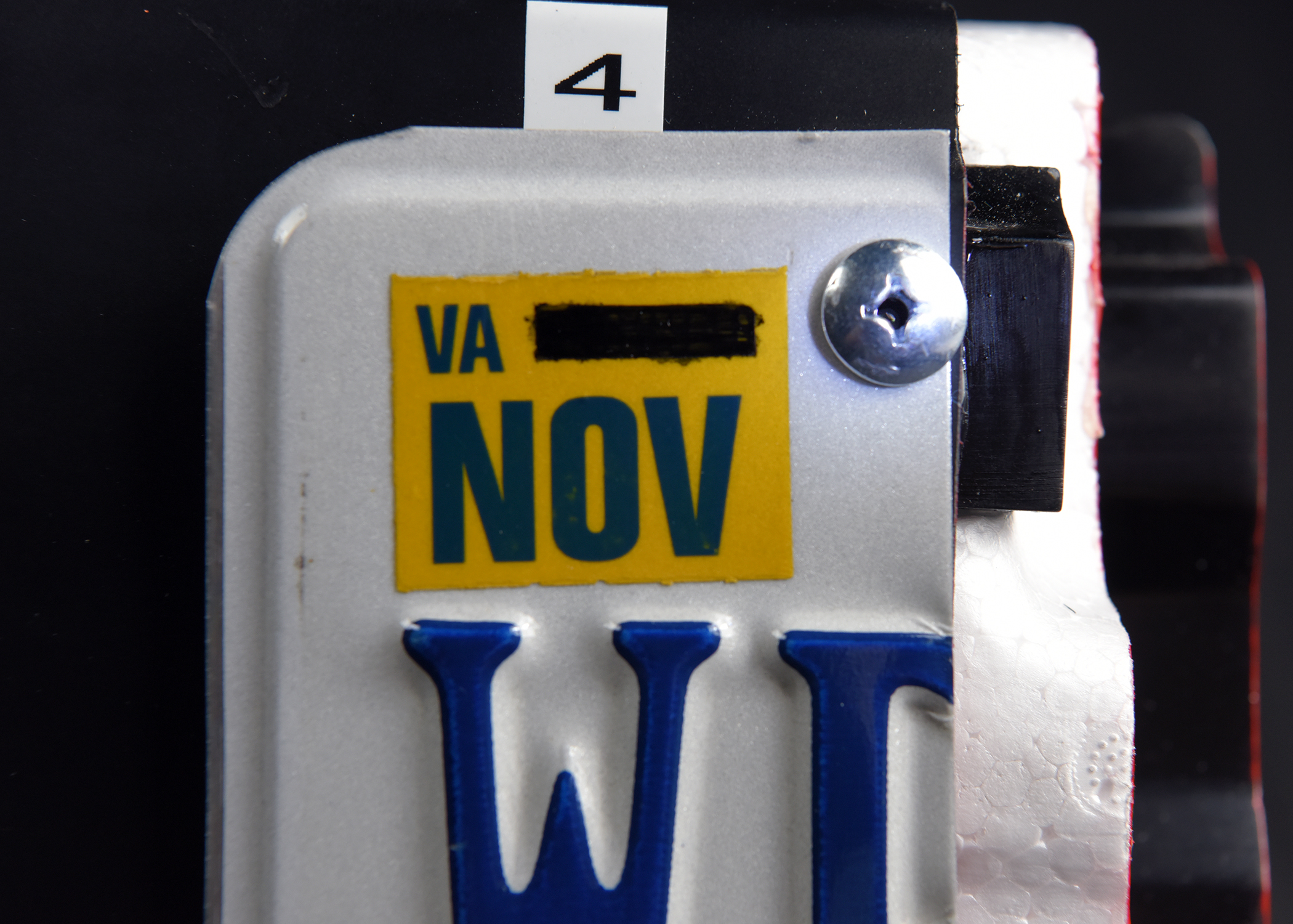 A close-up of the corner of a white license plate, showing a yellow Virginia (VA) sticker for November (NOV) and part of a blue letter 'W'. A small label with the number 4 is at the top.