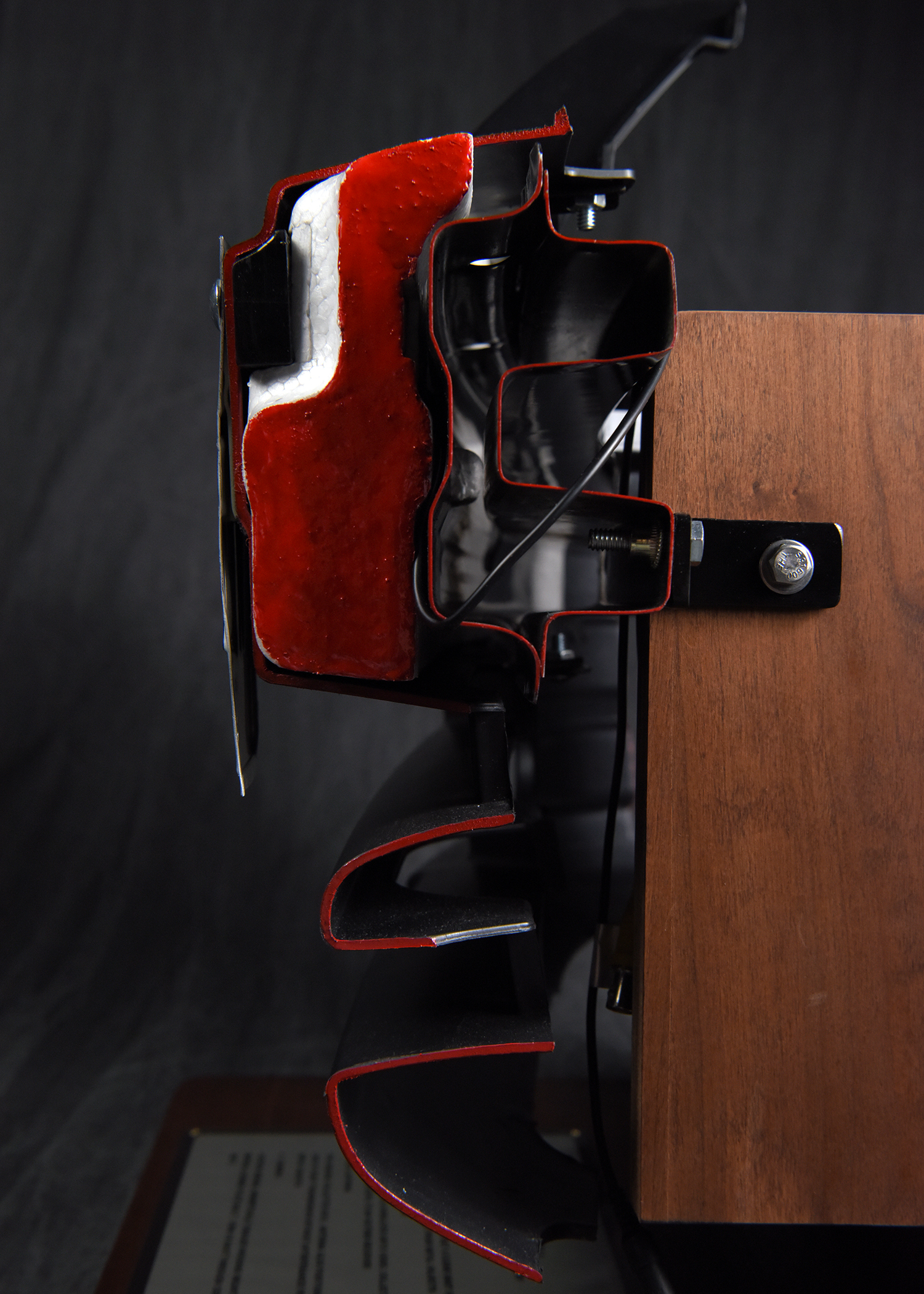 A close-up of a black metal latch mechanism attached to a glossy red and black object on the left, and bolted to a flat wooden surface on the right.