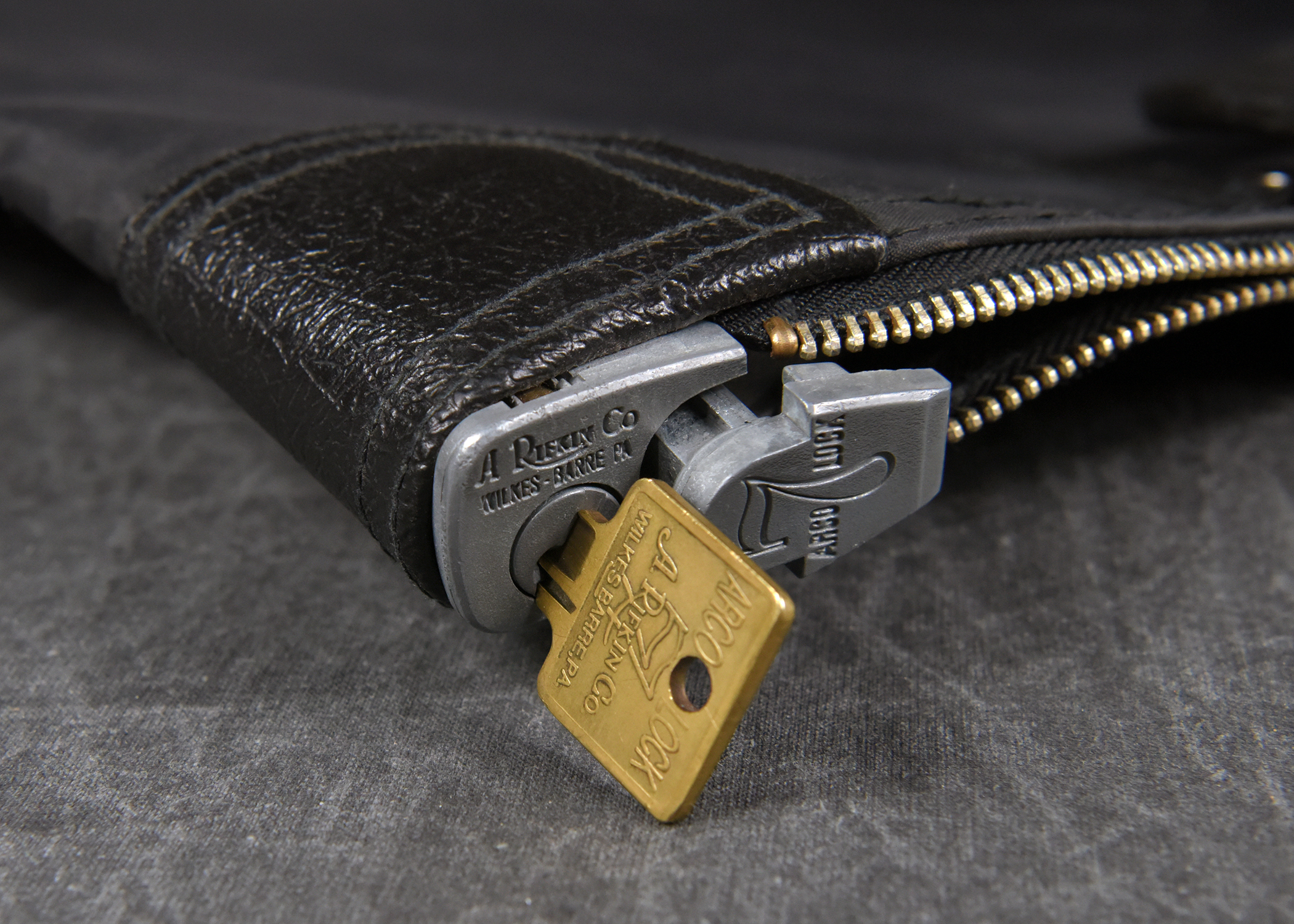 A close-up photograph of a silver zipper lock on a black leather bag. A small, square-headed brass key is inserted into the lock. The background is a dark gray fabric.