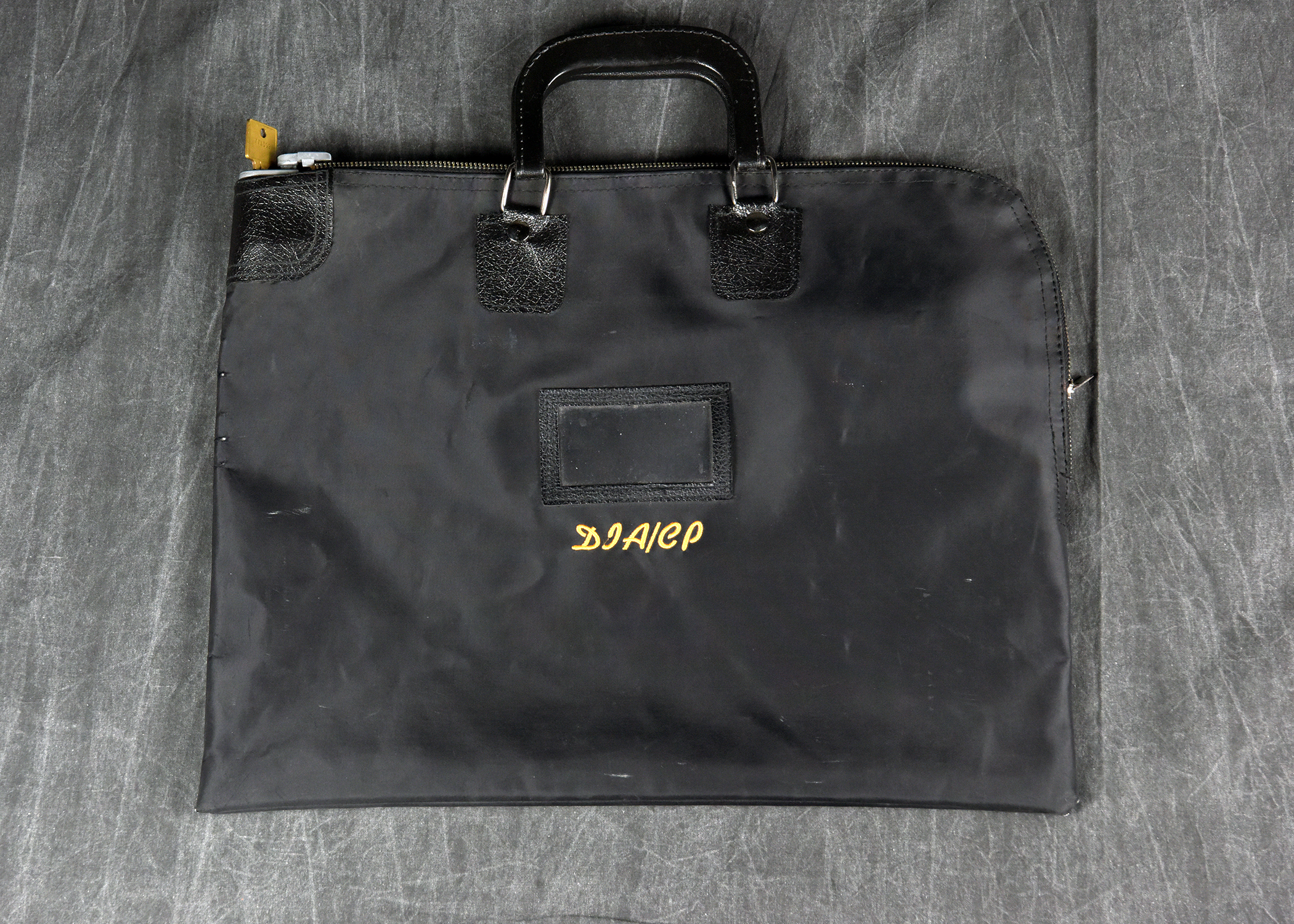 A black fabric document bag lying on a gray surface. The bag has a black handle, a zipper with a key lock, and the acronym 'DIACP' embroidered in gold thread on the front.