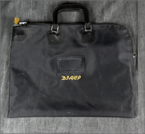 A black, zippered portfolio bag with leather handles and the letters 'DIA/CP' embroidered in yellow on the front. The bag is lying on a dark, rumpled fabric.