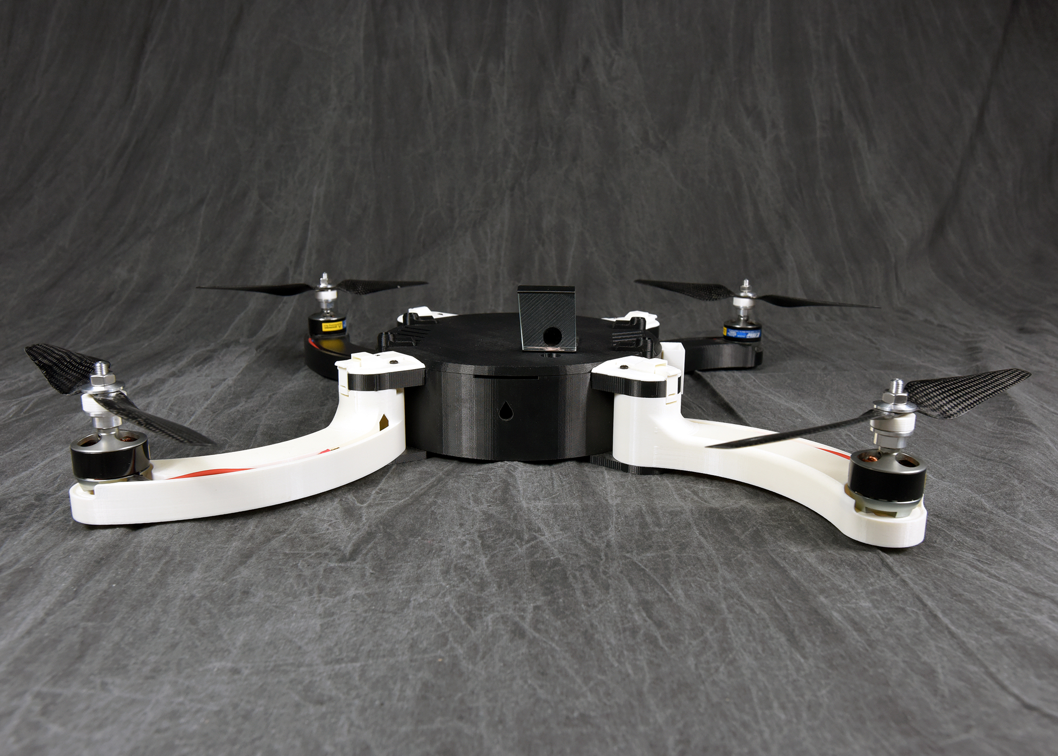 A front view of a fully assembled quadcopter drone. The drone has a black central body and four white arms extending outwards, each with a motor and a black propeller. It is sitting on a dark, wrinkled fabric background.