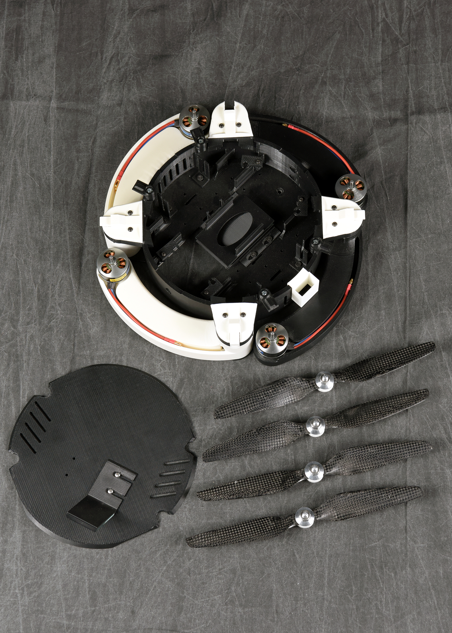 A top-down view of a disassembled drone. The main circular body sits in the center, with its white arms folded in. Beside it are the black top cover and four detached black propellers neatly arranged in a row.
