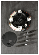 A top-down view of a disassembled quadcopter drone with its components arranged on a dark fabric background. The main body, with its arms folded, is shown alongside the four detached propellers and the top cover plate.