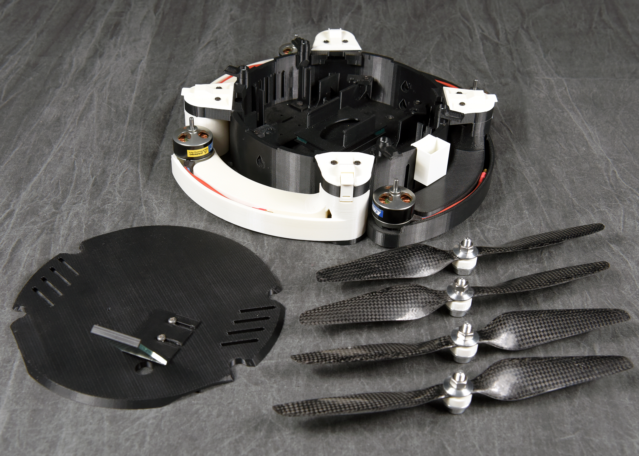 An angled view of a disassembled drone and its components. The main body, with white folded arms, is shown next to its black top cover and four detached black propellers, all resting on a dark, wrinkled fabric background.