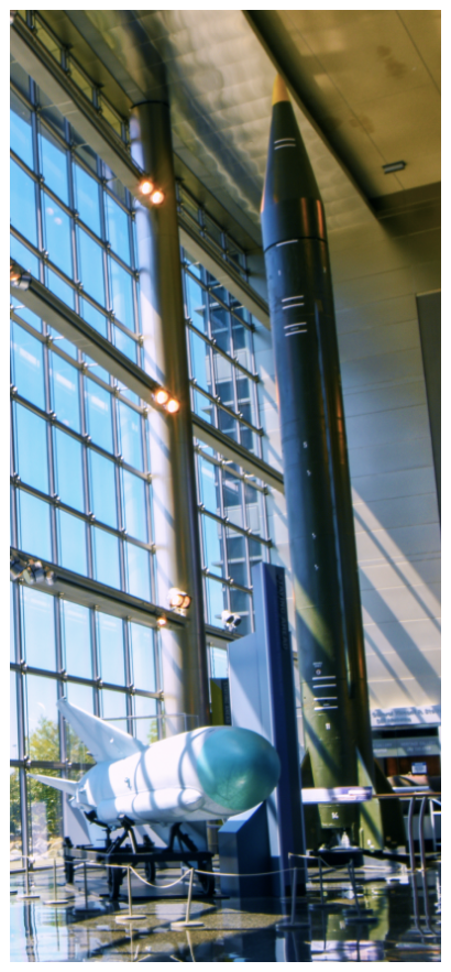 A tall, dark green Scud ballistic missile and a smaller, white surface-to-air missile displayed vertically inside a museum in front of a large, multi-story glass window.