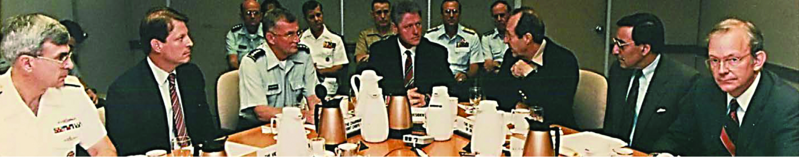 A panoramic photograph of a formal meeting, showing civilian leaders, including what appears to be former President Bill Clinton and Vice President Al Gore, seated at a conference table with military officials.