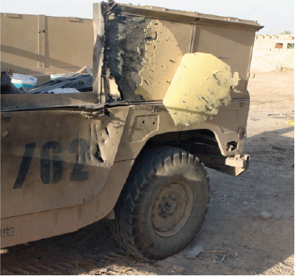 The rear quarter panel of a tan military vehicle, showing significant battle damage from shrapnel.