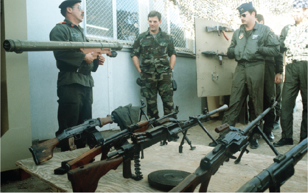 Several military personnel in different uniforms inspect a collection of captured weapons, including various machine guns and a rocket launcher, laid out on a table.